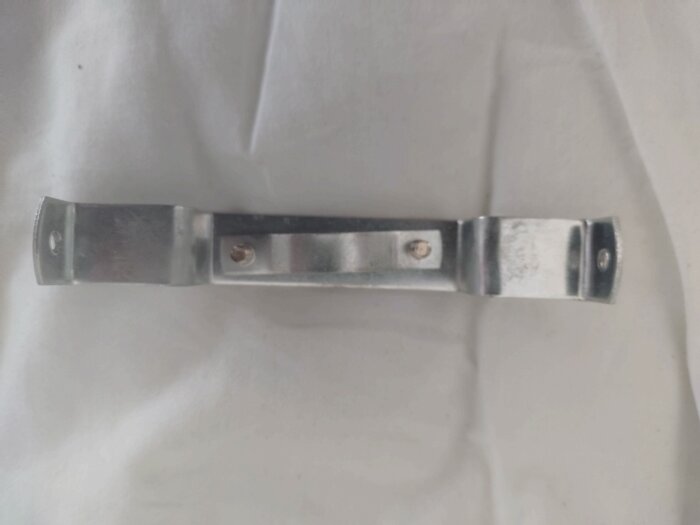 Metallbit med två skruvhål, troligen en del av ett lampsystem, ligger på vit yta.