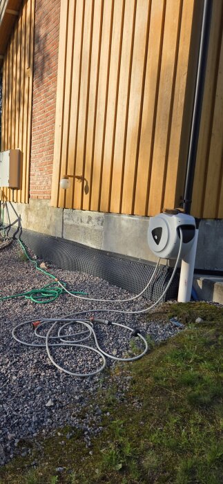 Fasadvägg med gul träpanel och röd tegel, svart plastlist löper längst botten; plastslang och kabelvindare i förgrunden på grus.