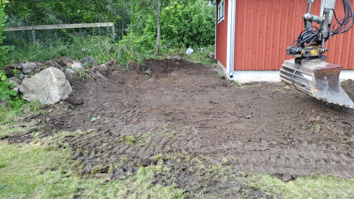 Grusplan vid ett rött hus med trädgårdsarbete, stenmur, grävskopa och grönt gräs. Förberett för utplaning och grässådd.