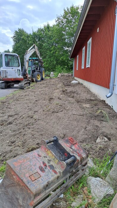 Grävmaskin vid en röd byggnad med omarbetad jord framför. Maskinredskap i förgrunden och träd i bakgrunden.