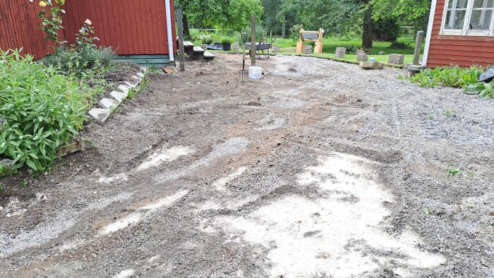 Grusyta med växter vid en röd byggnad, förberedd för rabatter och omfördelning av sten och jord, samt knäckstensmur.