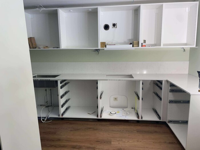 Renovering av kök med vita skåp och bänkskiva, utan fronter och diskho, med eluttag ovanför bänken och trä-imitationsgolv.