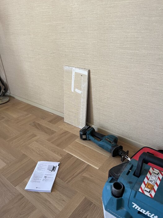 Gipsskiva och elektriskt verktyg på träparkettgolv framför beige tapet. En monteringsmanual ligger på golvet.