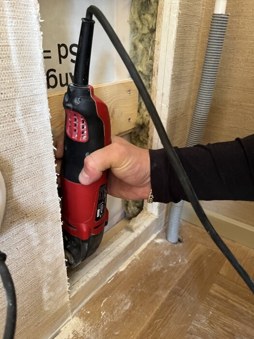En hand håller en röd elverktyg vid en träram med synlig isolering och gipsskiva, förbereder för finlir med gips och tapet.