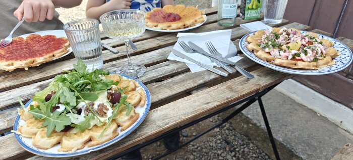 Tre tallrikar med våfflor på ett bord. En våffla med räkor och dill, en med sallad och en med sylt. Glas och bestick syns också.