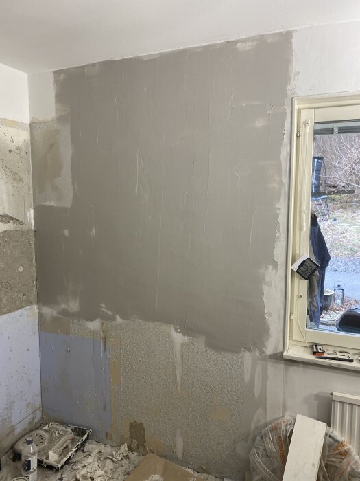 Betongvägg i renoveringsfas med ny spackling, nära fönster. Omgivande ytor är oordnade med verktyg och material på golvet.