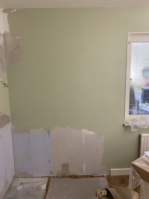 Grön vägg i renoveringsprojekt, delvis spacklad med olika lager gips och färg. Fönster till höger med plastskydd och golvklämmor synliga.