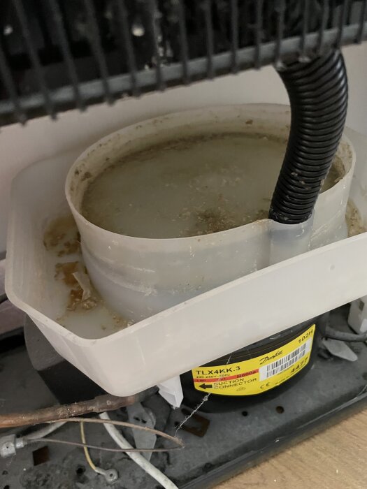 Vattenfylld droppskål bakom ett kylskåpsgaller med ett svart dräneringsrör kopplat till den.