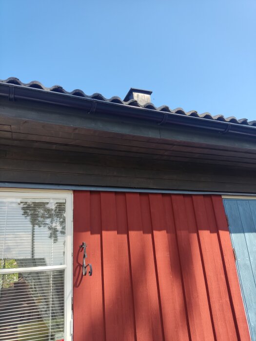 Takfot och vägg i en röd enplansvilla med tegeltak och vitmålat fönster, sedd från utsidan under klarblå himmel.