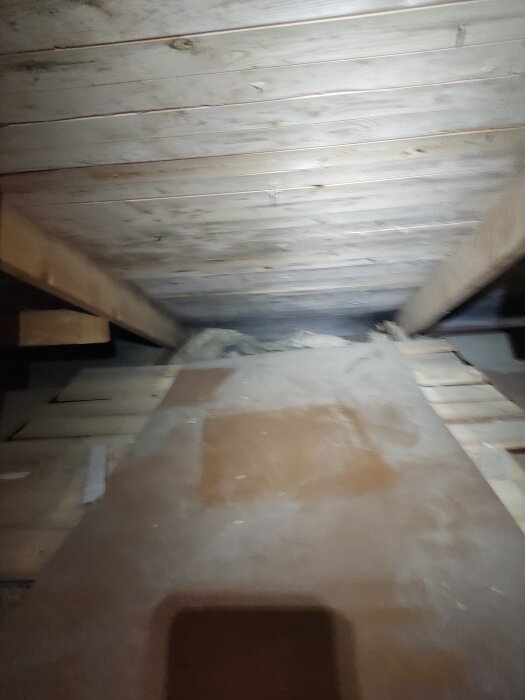 Loft med träbjälkar och isolering, sedd från vinden. Det finns damm och spån på ytorna och begränsat utrymme mot takfoten.