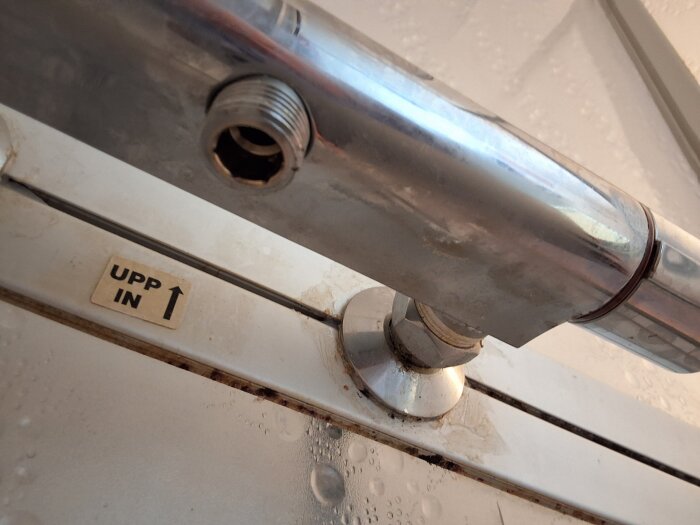 Utloppsrör från duschblandare med gängad anslutning och etikett med text 'UPP IN' och pil uppåt.