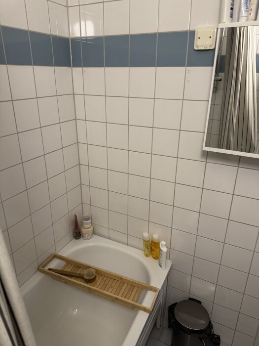 Badrum med vitt kakel och ett badkar med badkarshylla. Flaskor och borste synliga. Diskussion om att installera en tvättmaskin.