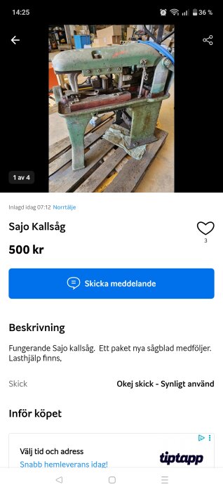 Gammal grön metallkallsåg på lastpall, synliga tecken på användning. Bakgrund består av lagerutrymme med kartonger och föremål.