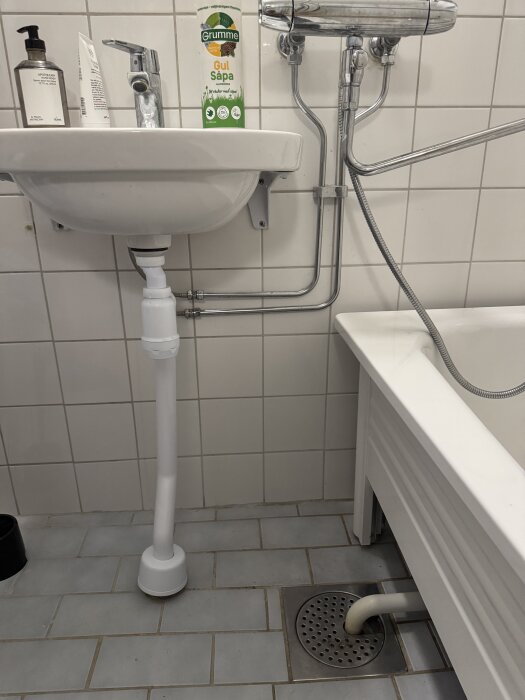 Badrum med handfat, kran, gul såpa, grå kakel, avloppsrör och badkar med duschmunstycke. Rör kopplade under handfatet och badkaret.