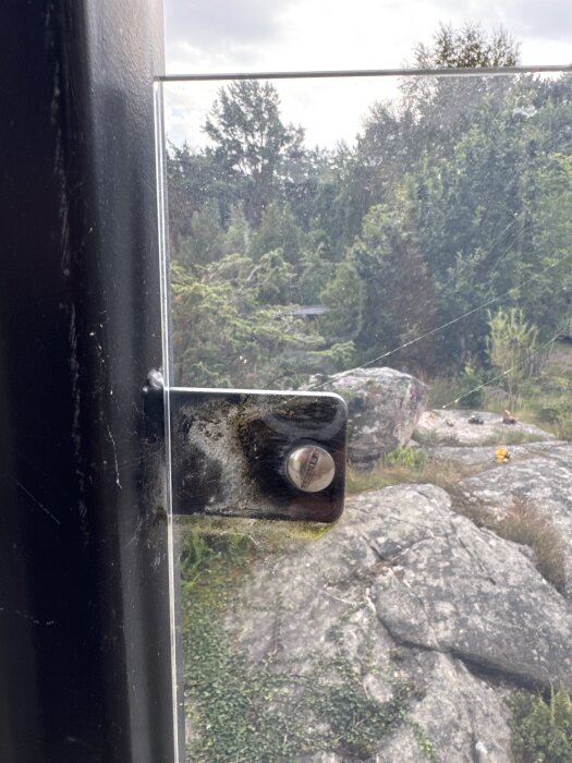 Balkongräcke i plast med metallfäste skruvat i en svart pelare, omgiven av träd och stenar i bakgrunden.