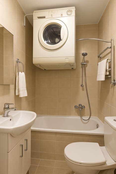 Badrum med tvättmaskin ovanför badkar, beige kakel, handfat och toalett. Förslag om glasvägg mellan maskin och dusch.