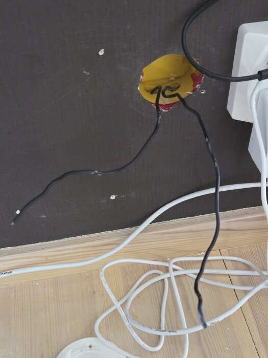 Två svarta kablar kommer ut ur ett gult hål i väggen, omgivna av vita kablar på ett trägolv.