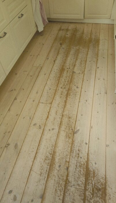 Vitbetsat furugolv med synligt slitage och mörka fläckar i kök eller matplats, typiskt efter åtta års användning.