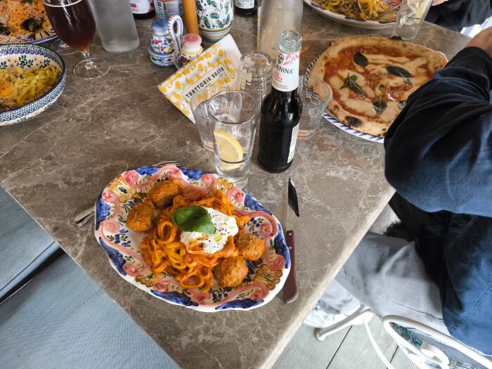 Flera tallrikar med olika pastarätter och en pizza på ett marmorbord, drycker och bestick syns också.