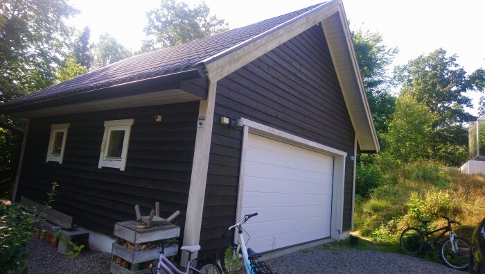 Ett mörkt trägarage med vita fönster och en vit port. Cyklar och vedstaplar syns framför garaget, samt träd och växtlighet i omgivningen.