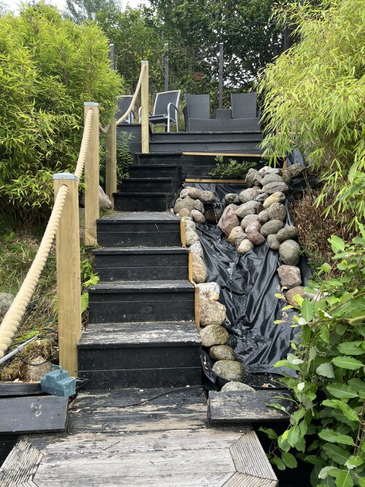 Trappa i trä i en trädgård, omgiven av buskar och stenar i en bäckfåra, med trädgårdsstolar i bakgrunden.