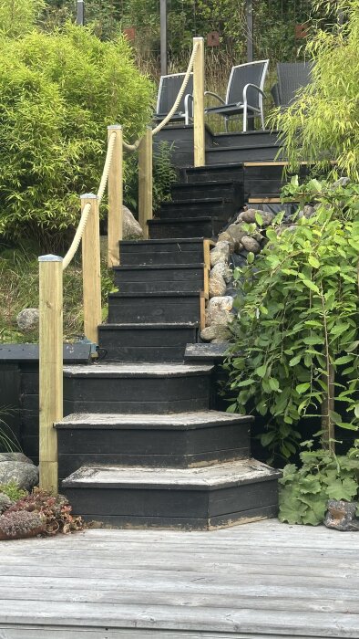 Svartmålad trätrappa med rep-ledstång, omgiven av grönska och stenar. Trappan leder upp till en sittplats med stolar.