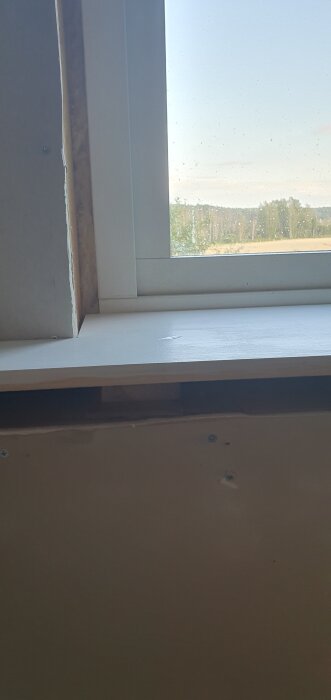 Fönsterbräda monterad vid ett fönster med synlig mellanrum mellan bräda och karm, med skog och himmel i bakgrunden.