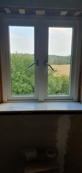 Fönsterbräda med öppningsbara fönster ovanför och utsikt över grönska och åker.