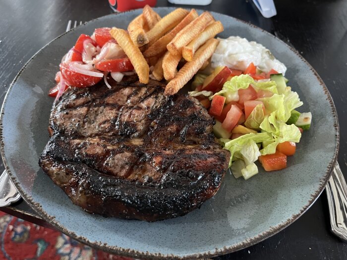 Grillad fläskkotlett med pommes, tomat- och löksallad, grönsallad och tzatziki på en tallrik.