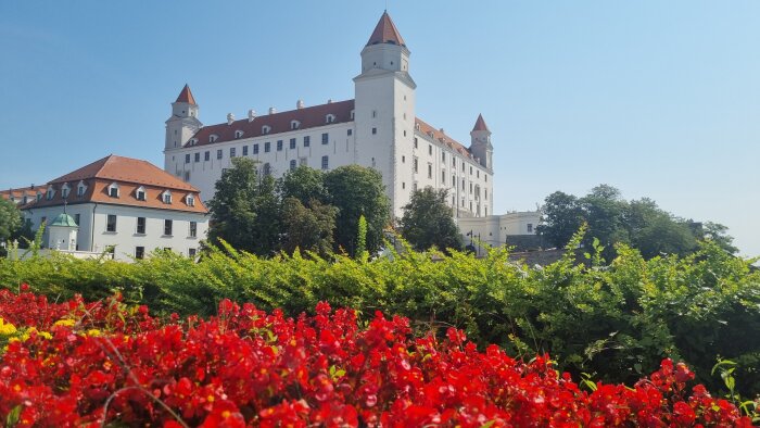 Vit byggnad med röda tak och tinnar, omgiven av grönska och röda blommor under en klarblå himmel.