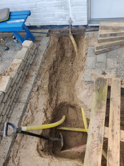 Grävt dike vid husvägg med synliga gula kablar och ett verktyg på marken, omgiven av stenar och trävirke.