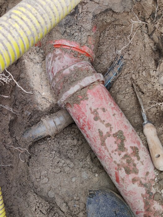 Grävt hål med synliga vatten- och avloppsrör, inklusive en röd anslutning och blått rör, i samband med renovering av vattenledning.