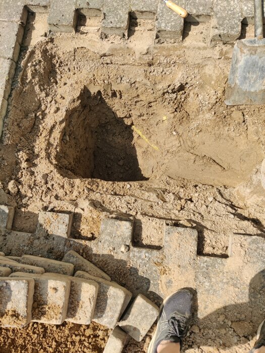 En nygrävd grop med runt form, omgiven av flyttade stenplattor, en spade syns till höger, samt kanten av en sko längst ner i bilden.