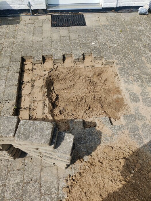 Uppgrävd ruta i stenbelagd uteplats med borttagna stenplattor lagda åt sidan, synlig sand och jord för rörarbete.