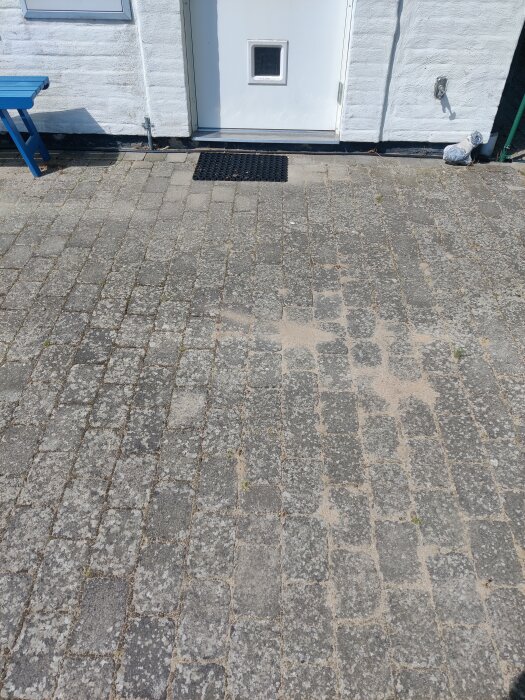 Dörr in i ett hus med vit vägg och stenlagd yta framför. Plattorna visar spår av nyligen utfört arbete och grävning.