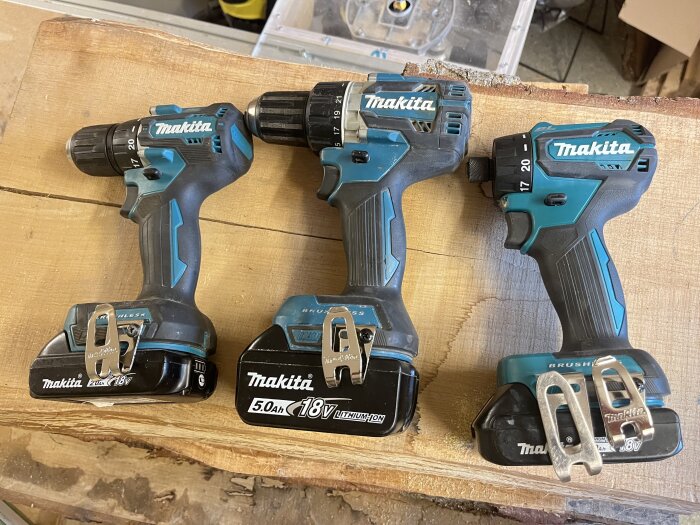 Tre Makita borrmaskiner med bälteskrokar och olika batteristorlekar på ett träbord. Två maskiner med 18V och en med 12V.