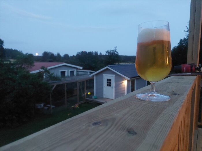Ett ölglas på ett träräcke vid skymningen med hus i bakgrunden.