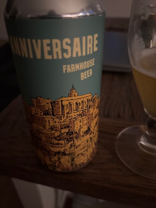 En burk med "ANNIVERSAIRE FARMHOUSE BEER" visas bredvid ett glas öl på ett bord.