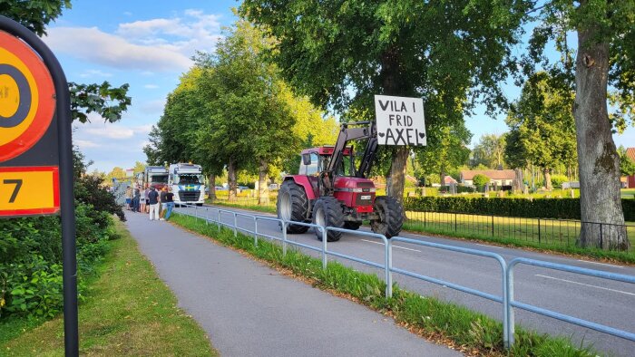 Traktor med skylt "Vila i frid Axel" i kortege på landsväg, med träd och hus i bakgrunden.