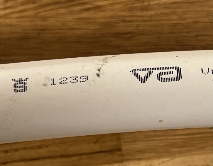 Vit rörsektion med tryckt text och vissa avskavda delar, tydlig text är "72 1.2339" med symboler. Bakgrund i trä med fokus på rördetaljer.