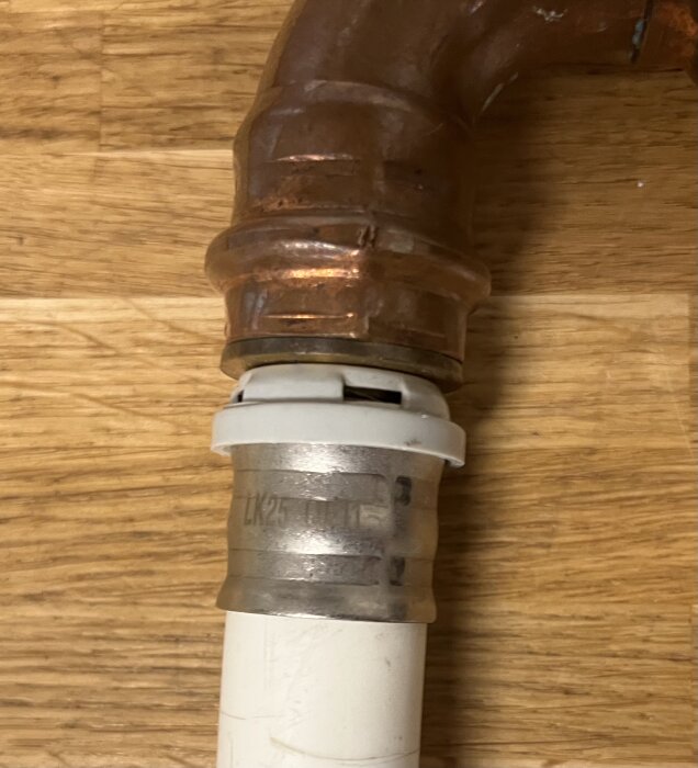 Kopparrör anslutet till en vit plastledning via en metallkoppling märkt "LK25 QFIT", på ett träunderlag.