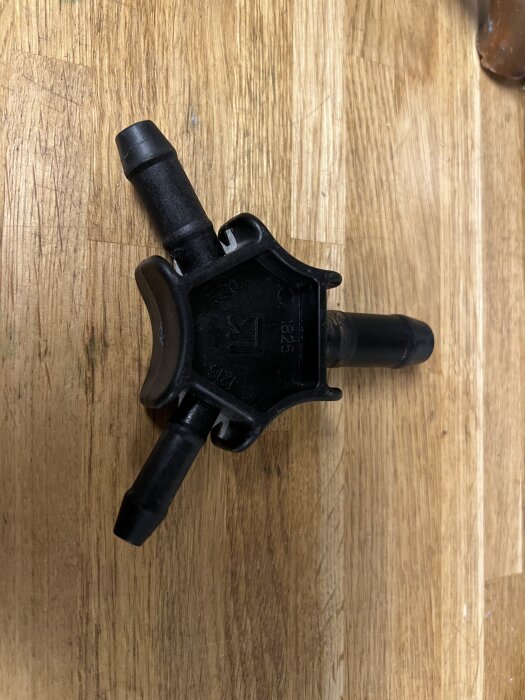 Svart plastkoppling med tre anslutningar, avsedd för rör, placerad på en träyta. En del av värmesystemets komponenter.