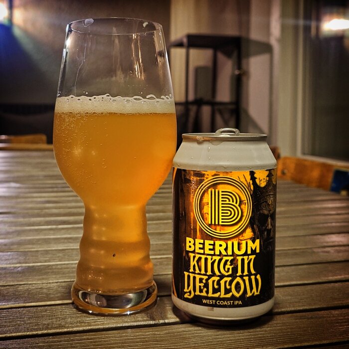 En ölburk märkt "Beerium King in Yellow West Coast IPA" vid sidan av ett glas med öl på ett träbord.