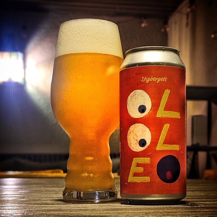 Ett glas öl bredvid en ölburk med röd etikett, texten "Olle" och abstrakt design på bordet i en dämpad miljö.