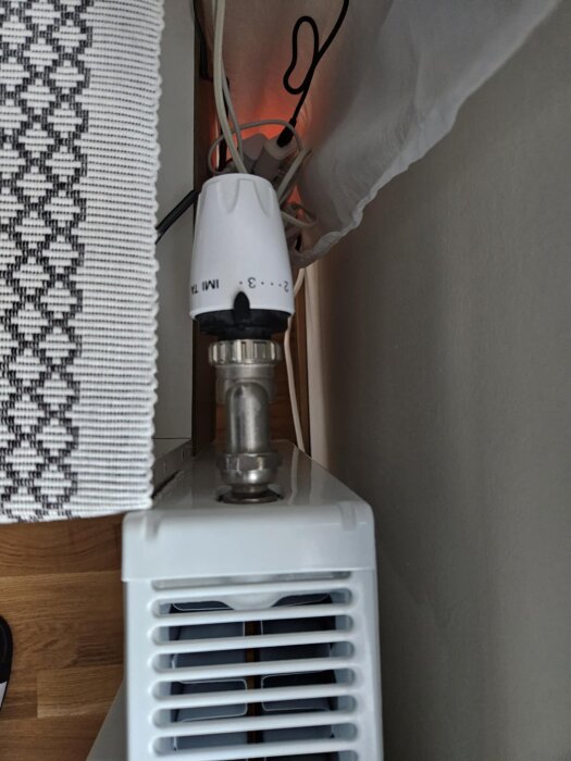 Radiator med vit termostat och anslutna kablar i närbild, monterad intill en vägg; fråga om byte av termostatens placering.