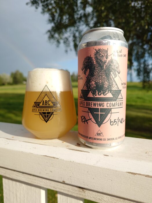 Burk och glas av Apex Brewing Company Phantom Current IPA på ett räcke, med grönska och regnbåge i bakgrunden.