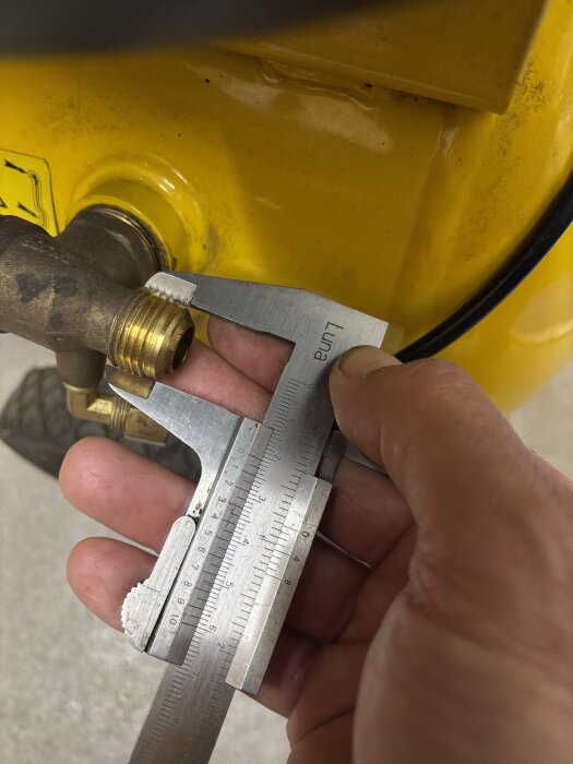 Hand håller skjutmått mot en gängad anslutning på en gul kompressor för att mäta kopplingen.