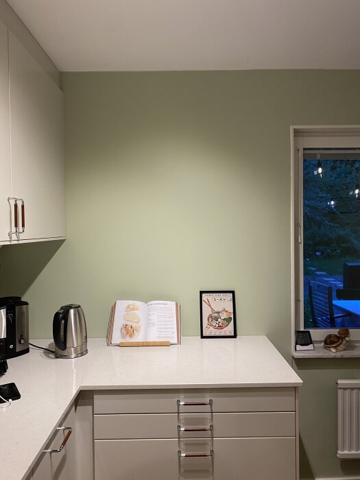 Grönmålad köksvägg med vit bänkskiva, tekokare, uppslagen kokbok och ram. Fönster med utsikt över trädgård i bakgrunden.