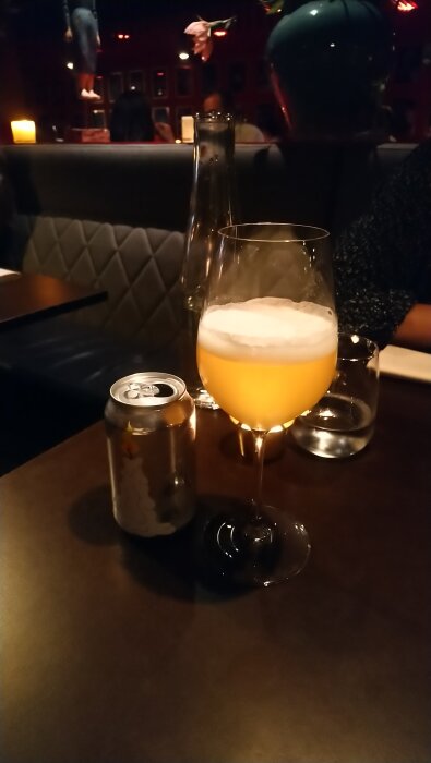 Ett glas med alkoholfri Oat Pale Ale och en läskburk, stående på ett bord i en dunkel miljö med ljus och flaskor i bakgrunden.