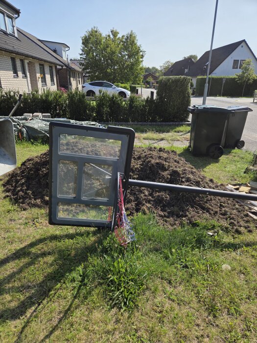 Hög med uppgrävd jord på en gräsmatta, med en omkullvält basketkorg i förgrunden och hus i bakgrunden.